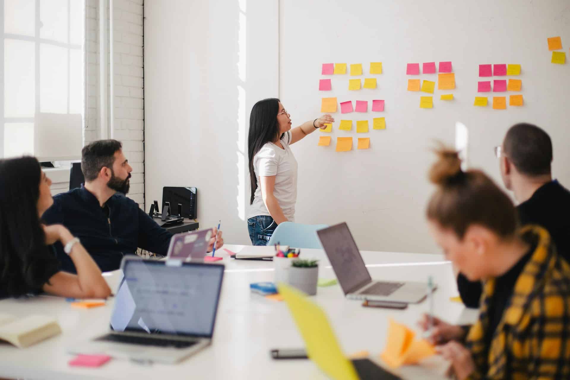 Mehrere Personen sitzen vor ihren Laptops an einem Konferenztische und arbeiten und unterhalten sich. Eine Frau steht vorne und zeigt auf ein Whiteboard