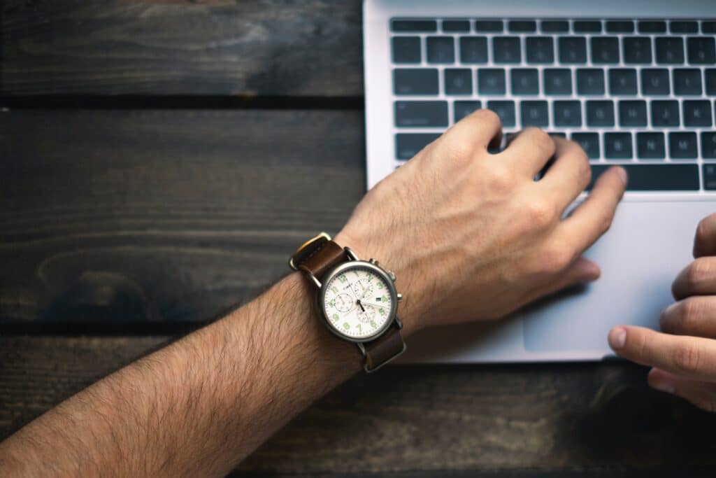Man sieht einen Tisch aus der Vogelperspektive. Darauf steht ein Laptop, bei dem Bildausschnitt ist allerdings nur die Tastatur zu sehen. Auf der linken Seite sieht man den Arm einer Person mit einer braunen Armbanduhr am Handgelenk