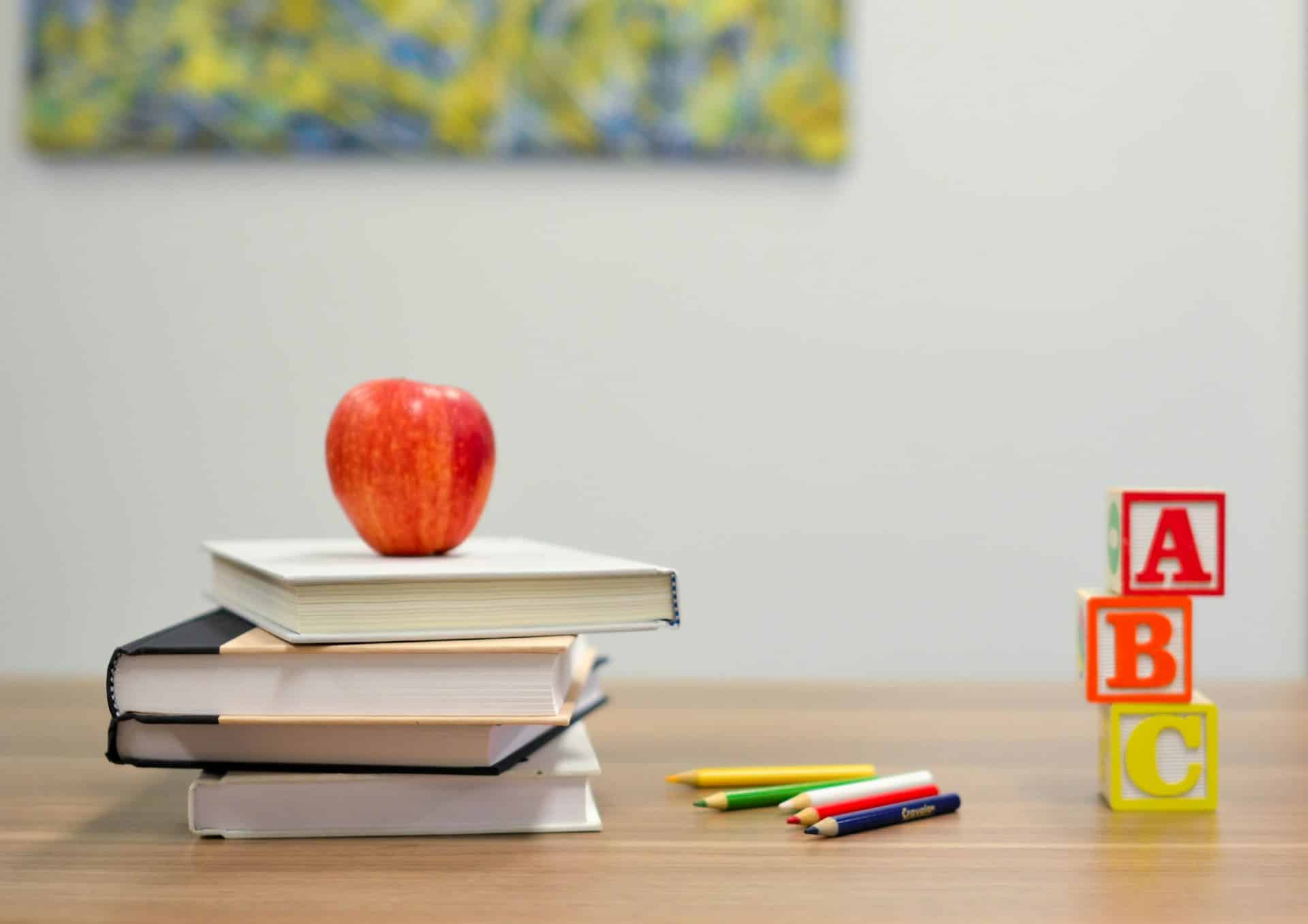 Auf einem Tisch liegen vier Bücher aufeinander gestapelt. Darauf liegt ein Apfel. Links daneben liegen mehrere verschiedenfarbige Stifte. Neben den Stiften stehen drei Holzblöcke mit den Buchstaben A, B und C.