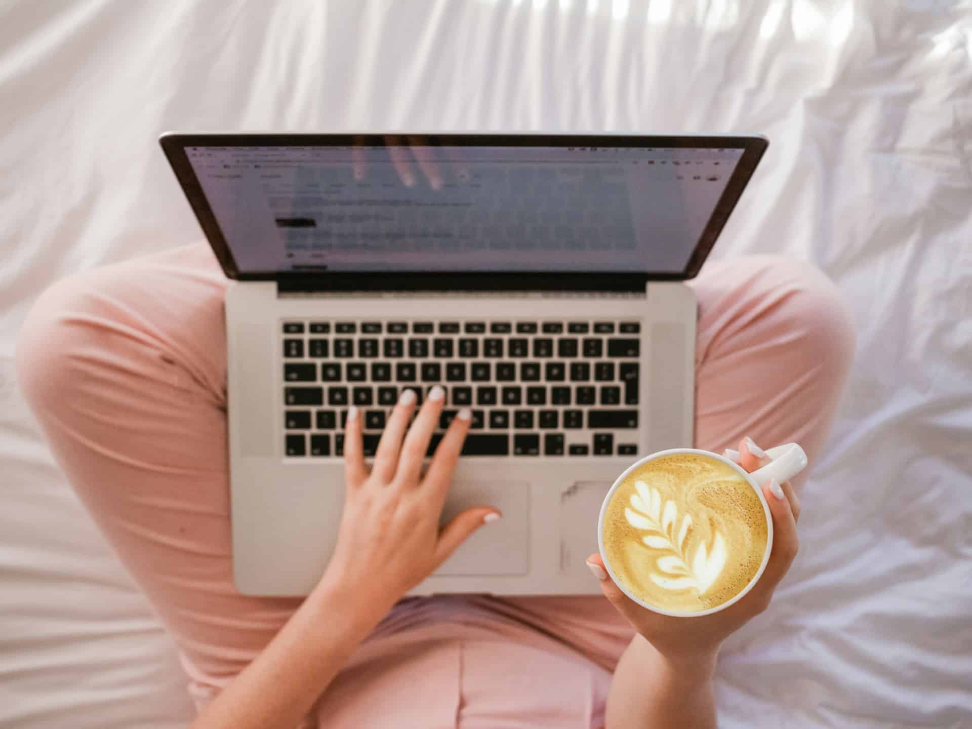 Eine Person sitzt auf mit dem Laptop auf dem Bett und und hält eine Kaffeetasse in der rechten Hand. Das Bild ist aus der Vorgelperspektive aufgenommen
