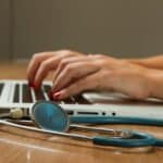 Eine Frau tippt an einem Laptop, davor liegt ein Stethoskop