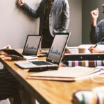 Working Out Loud: Die Methode für bessere Vernetzung An einem Konferenztisch sitzen mehrere Personen, alle haben einen Laptop vor sich stehen. Eine Person steht vor Kopf an einem Whiteboard und erklärt etwas
