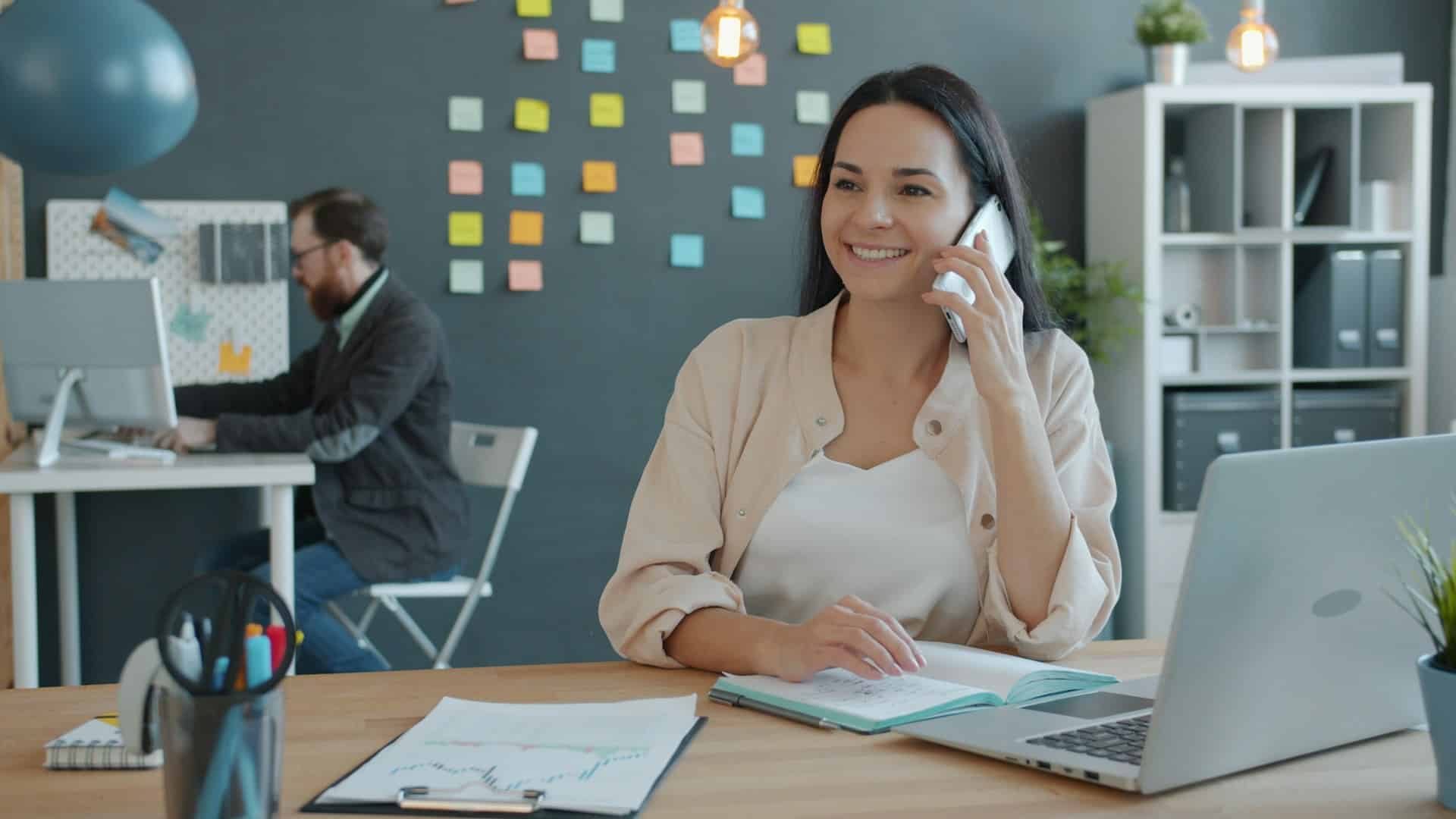 Eine Frau sitz am Schreibtisch vor einem Laptop und telefoniert. Im Hintergrund ist ein weitere Schreibtisch mit einer Person zu sehen.